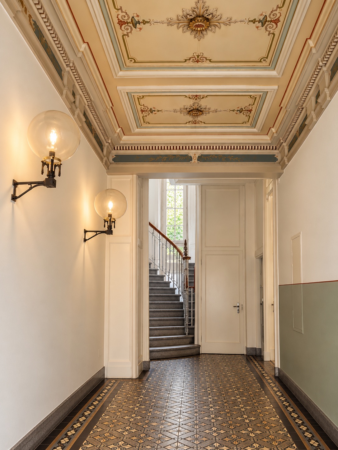 An entrance hall with a painted ceiling, globe lamps and a curving staircase beyond